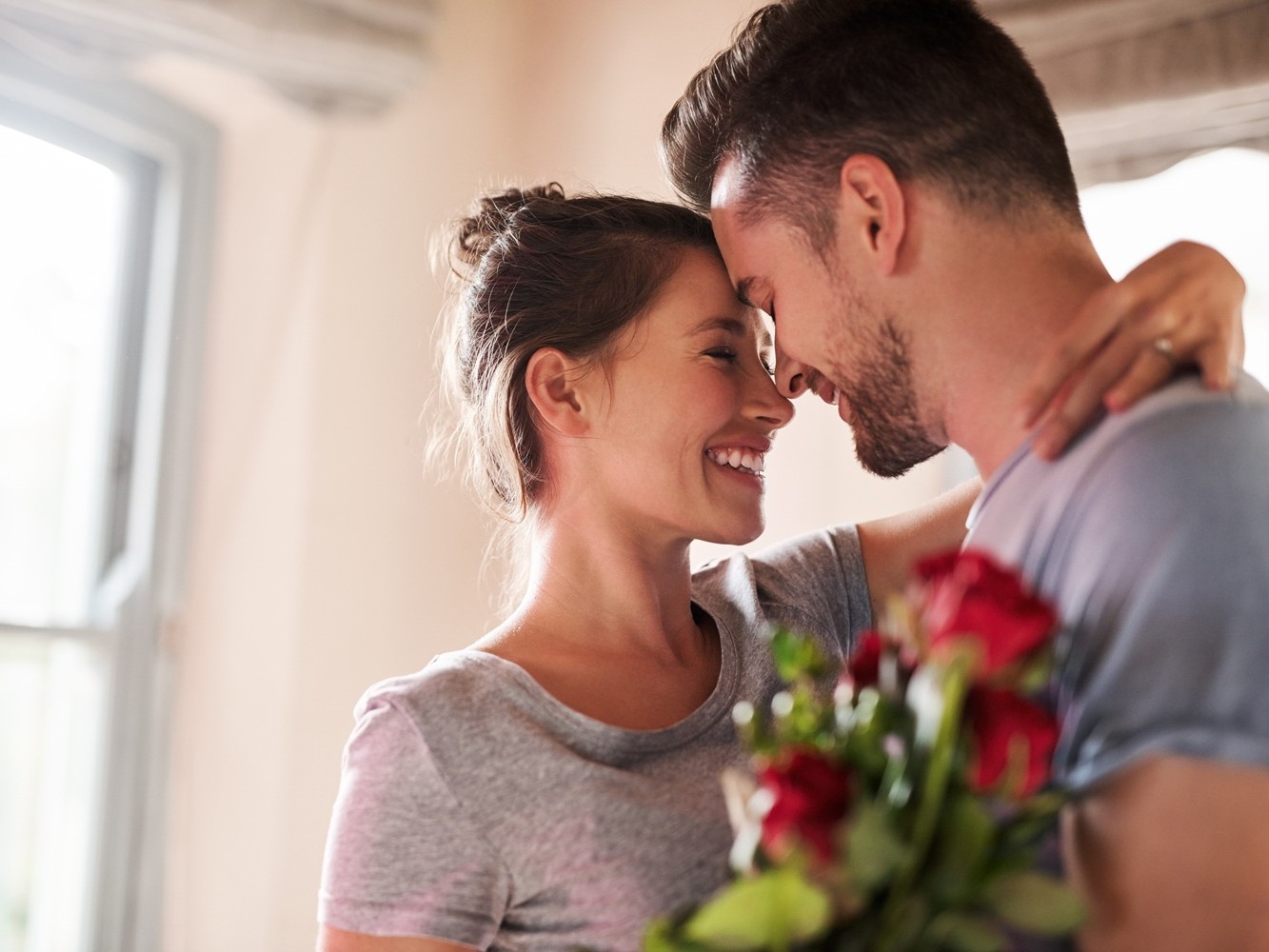 Flowers, happy couple and present in home for love, connection and bonding together with hug. Affection, man and woman with red roses for relationship, commitment and anniversary celebration in house